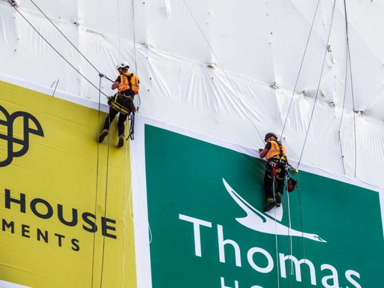 Rope access technicians installing banner on to scaffolding sub frame on shrink-wrap